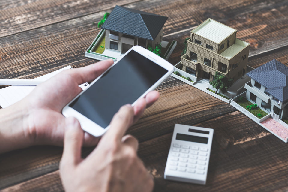 Eine Person rechnet vor drei Miniaturmodellen von Einfamilienhäusern mit dem Smartphone etwas aus