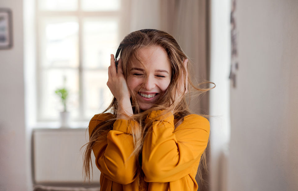 Eine junge Frau mit Kopfhörern hört in ihrem Zimmer vergnügt Musik und lacht