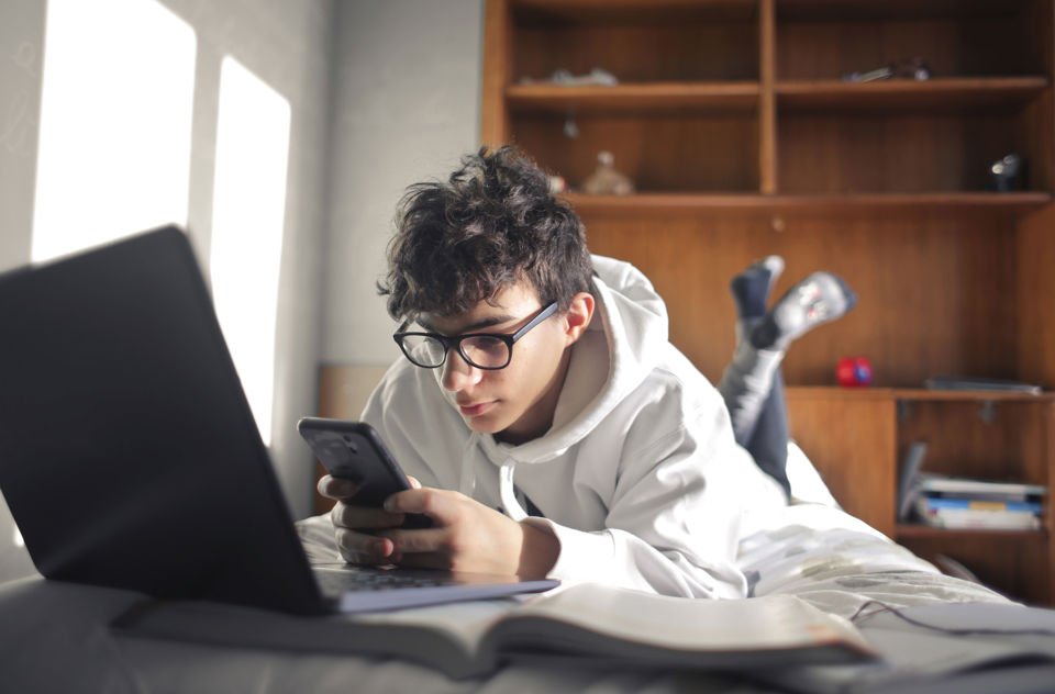 Ein Jugendlicher liegt mit dem Notebook auf dem Bett und benutzt sein Smartphone