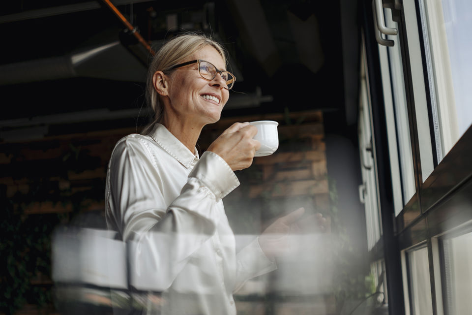 Eine Frau im Pensionsalter blickt entspannt mit einer Tasse in der Hand aus dem Fenster