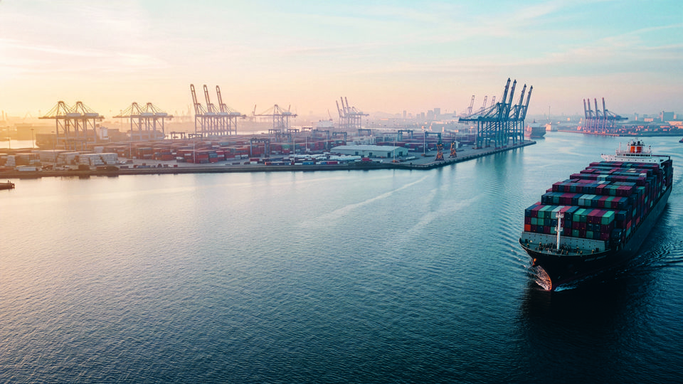 Containerschiff auf einem Fluss in der Nähe eines Hafens: Titelbild des Anlagemagazins /sicht herausgegeben von der St. Galler Kantonalbank, Ausgabe Oktober 2026 zum Thema "Das diskrete Rückgrat der Weltwirtschaft"