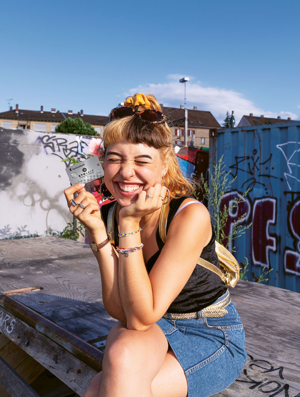Aufgeweckte Teenagerin sitzt in urbaner Umgebung auf einem Absatz und zeigt lächelnd ihre StuCard